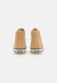 Tan suede vysoké tenisky s texturovaným povrchem, černou a bílou gumovou podrážkou a kruhovým logem na vnější straně.