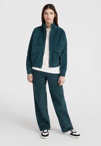 Giacca in velluto a coste verde acqua con due tasche frontali, polsini e orlo elastici, abbinata a pantaloni coordinati. T-shirt bianca sotto, sneakers in bianco e nero.