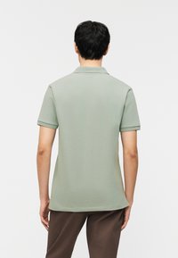 Άνδρας που φοράει ανοιχτό πράσινο πόλο με κοντά μανίκια και σκούρο καφέ παντελόνι, στέκεται γυρισμένος ανάποδα σε απλό ανοιχτό φόντο.