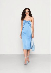 Vestido midi de cetim azul com detalhe em laço, alças finas ajustáveis, fenda lateral, combinado com saltos altos e uma carteira a condizer.