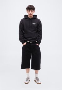 Νεαρός με σγουρά μαλλιά στέκεται με τα χέρια στις τσέπες φορώντας μαύρο φούτερ Levi's, μαύρες φαρδιές βερμούδες, λευκές κάλτσες και ασημένια αθλητικά παπούτσια σε λευκό φόντο.