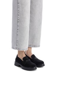 Mocasines de ante negro con una suela de goma gruesa y un acento metálico. Combinados con jeans de pierna ancha recortados en gris claro.