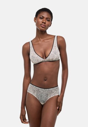 Femme debout, portant un soutien-gorge et une culotte assortis à imprimé floral beige avec bordure noire, sur un fond uni gris clair.