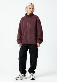 Veste en polaire bordeaux avec surface texturée, col montant, fermeture éclair et poches avant ; associée à un pantalon cargo noir et des baskets.