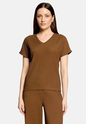 Femme aux cheveux lisses et foncés, portant un haut marron à manches courtes en V et un pantalon assorti, debout devant un fond clair uni.