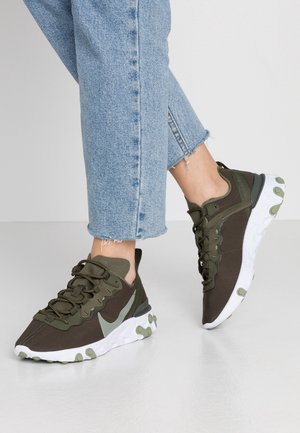 Osoba nosząca jasnoniebieskie jeansy typu cropped i ciemnozielone trampki z białymi podeszwami, stojąca na jasnoszarej podłodze.