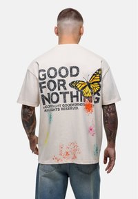 Beżowy bawełniany t-shirt z dużym czarnym napisem "NIE PRZYDATNY", grafiką żółtego motyla oraz kolorowymi akcentami w formie plam na plecach.