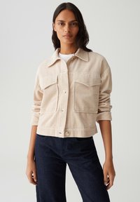 Beige Kordsakko mit zwei Brusttaschen, Knopfverschluss und hochgekrempelten Ärmeln, getragen über einem weißen Top und kombiniert mit dunklen Jeans.