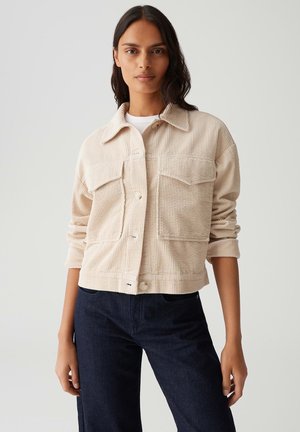 Veste en velours côtelé beige avec deux poches poitrine, fermeture à boutons et manches retroussées, portée sur un haut blanc et associée à un jean sombre.
