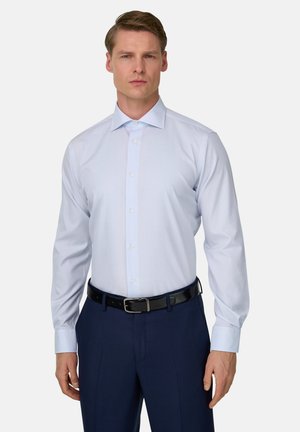Homme portant une chemise bleue claire rentrée dans un pantalon bleu marine avec une ceinture noire, se tenant contre un fond blanc uni.