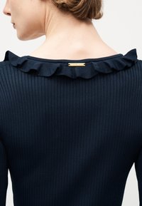 Πλάτη γυναίκας που φοράει σκούρο μπλε ριμπ τοπ με βολάν στο γιακά και μικρή χρυσή ετικέτα Michael Kors στο λαιμό.