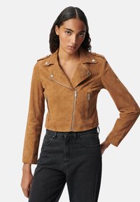 Joven con cabello largo y rizado oscuro, vistiendo una chaqueta de ante marrón corta y jeans negros de cintura alta, de pie con una mano en la cadera.