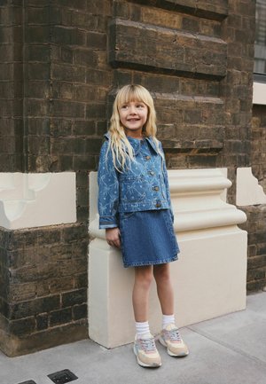 Mladé dievča s dlhými blond vlasmi sa opiera o tehlovú stenu, oblečená v vzorovanej džínsovej bunde, džínsovej sukni, bielych ponožkách a pastelových teniskách.