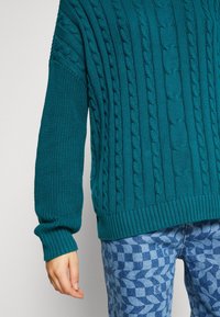 Miętowy dziergany sweter z fakturą warkocza, ściągaczami na rękawach i u dołu. Połączony z niebieskimi dżinsami we wzory o geometrycznym motywie.