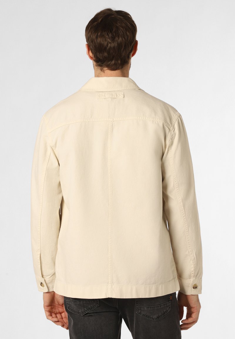 Hombre vestido con una chaqueta beige con puños abotonados, de pie de espaldas, combinado con jeans gris oscuro contra un fondo sencillo.