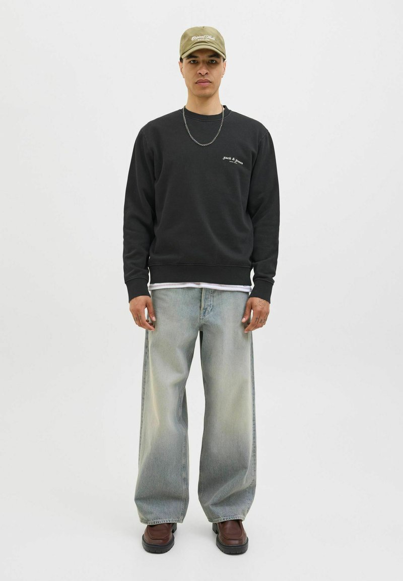 Black sweatshirt with white embroidery, loose grey jeans, and brown shoes. Worn with a light green cap and silver chain necklace.