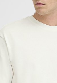 Svetlo béžová mikina s okrúhlym výstrihom, jemnou textúrou a minimalistickým dizajnom. Detailný pohľad zdôrazňuje hladký povrch textílie.
