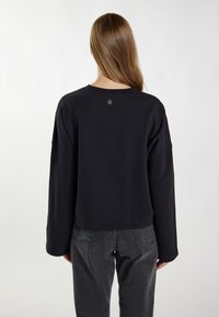 Schwarzes, cropped Sweatshirt mit langen Ärmeln, gerippter Textur und rundem Halsausschnitt. Besitzt ein Logo-Label am Rücken in der Nähe des Halses.