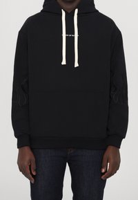 Sudadera negra con cordones blancos, pequeño texto blanco en el pecho y sutiles diseños de llamas en las mangas, combinada con jeans azul oscuro.
