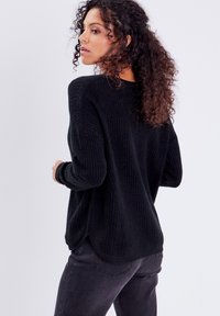 Schwarzer gestrickter Pullover mit lockerer Passform, der ein geripptes Muster, lange Ärmel und einen leicht abgerundeten Saum aufweist. Kombiniert mit dunklen Jeans.