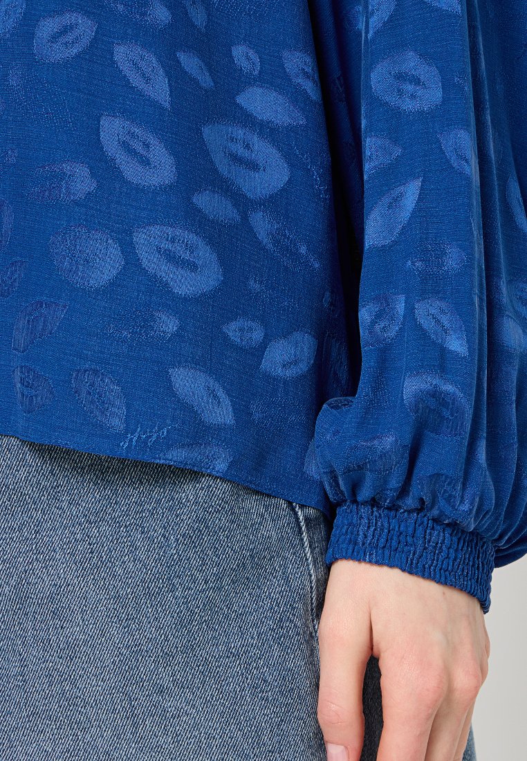 Blaue Bluse mit einem strukturierten Muster, mit lockeren Ärmeln und einem smockierten Bund. Kombiniert mit hellem Denim für einen Kontrast.