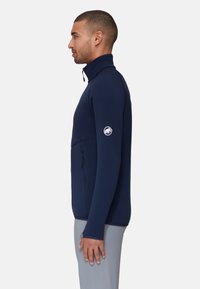 Marineblauwe fleece zip-up jas met hoge kraag, zijzak met rits en logopatch op de mouw. Gladde textuur en getailleerd ontwerp.
