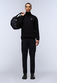 Zwarte turtleneck trui met rond logo, zwarte cargobroek met zijzakken, en zwarte schoenen, terwijl hij een zwarte duffeltas vasthoudt.