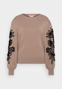 Pull beige avec des motifs floraux noirs brodés sur les manches. Présente des poignets côtelés et un col rond en tissu tricoté doux.