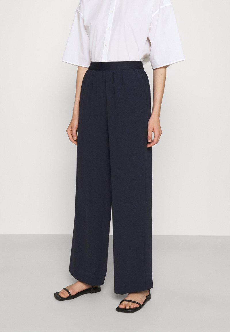 Personne portant un pantalon bleu marine taille haute à jambes larges, une chemise blanche à manches courtes boutonnée et des sandales plates noires, debout devant un fond uni.