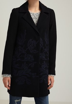 Manteau noir avec manches texturées et broderie florale subtile porté sur un pull en maille gris clair et un jean bleu déchiré.