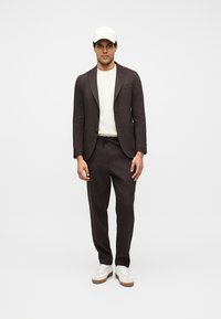 Homme portant un costume marron foncé avec un pantalon à cordon, un t-shirt blanc, une casquette de baseball blanche et des baskets blanches, debout devant un fond uni.