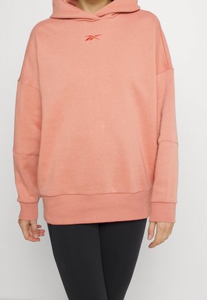 Osoba ubrana w luźną koralową bluzę z kapturem z małym czerwonym logo Reebok i czarne legginsy, stojąca na jednolitym tle.