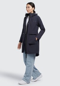 Giacca impermeabile blu marina con cappuccio, zip frontale e due tasche con zip. Presenta una vita stretta e una texture del tessuto liscia.