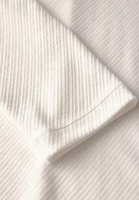Witte ribbelstof met een zachte textuur, die een gedeelte van een mouw toont met een nette zoom en subtiele stiksels.