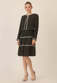 Traje de tweed negro con chaqueta ajustada y falda con tablas. Presenta detalles en perlas blancas y un diseño estructurado. Incluye tacones plateados con tiras.