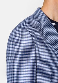 Marynarka o teksturowanym granatowo-białym wzorze w kratę, z wyraźnym kołnierzem i dopasowanym ramieniem, podkreślająca formalne elementy wzornictwa.
