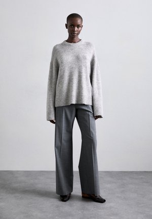 Gai pelēks pārāk liels adīts džemperis ar ribotu apakšmalu, saskaņots ar pielāgotām pelēkām platām biksēm un atvērtu melno apavu. Gludas tekstūras.