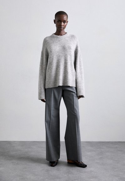 Pulover supradimensionat de tricot gri deschis, cu tiv gulos, asortat cu pantaloni largi gri, tăiați, și pantofi negri cu vârf deschis. Texturi netede.