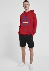 Röd hoodie med blå och vit logotypkontrast, svarta shorts och vita sneakers. Hoodien har en kängurulomme och dragsko på huvan.