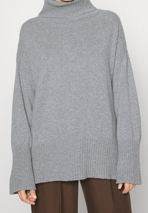 Lässiger grauer Rollkragenpullover mit gerippten Bündchen und Saum, getragen zu braunen Hosen.