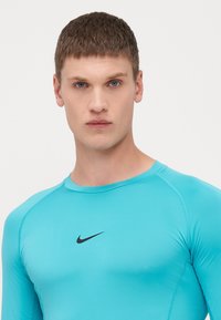Lys blå sportsbluse med lange ærmer, lavet af strækbart materiale. Har et sort Nike-logo på brystet. Glat tekstur og tætsiddende design.