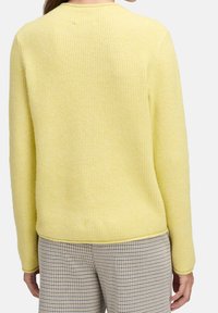 Vue de dos d'une personne portant un pull en maille côtelée jaune pâle et un pantalon à carreaux beige et marron, debout les bras détendus le long du corps.