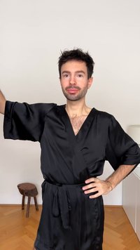 Juodas satino chalatas su plačiomis rankovėmis ir diržo ryšiu, pasižymintis lygiu audiniu ir giliais iškirpte. Šviesaus medžio taburetė fone.