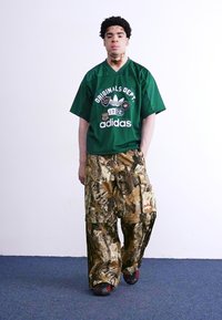 Zaļš lielizmēra Adidas krekls ar logo un plankumiem. Brīva piegriezuma kamuflāžas krāsu cargo bikses. Melna apavi ar sarkaniem akcentiem. Viens pats fons.