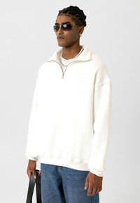 Witte zip-up sweatshirt met hoge kraag, verlaagde schouders en geribbelde manchetten. Gecombineerd met blauwe jeans en accessoires, waaronder een zonnebril.