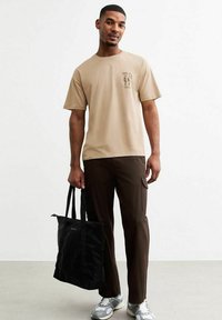 Camiseta de algodón beige con diseño gráfico, combinada con pantalones cargo marrones y una bolsa tote negra. Los zapatos son grises con acentos claros.