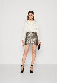 Langärmliges cremfarbenes Oberteil mit drapierter Halslinie, kombiniert mit einem taillierten Minirock aus silbernen Pailletten. Schwarze Absätze und eine Clutch vervollständigen das Outfit.