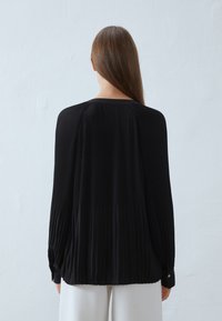 Blouse plissée noire avec des manches longues, un col rond et des poignets boutonnés. Elle dispose d'un tissu texturé et d'une silhouette ample et fluide.
