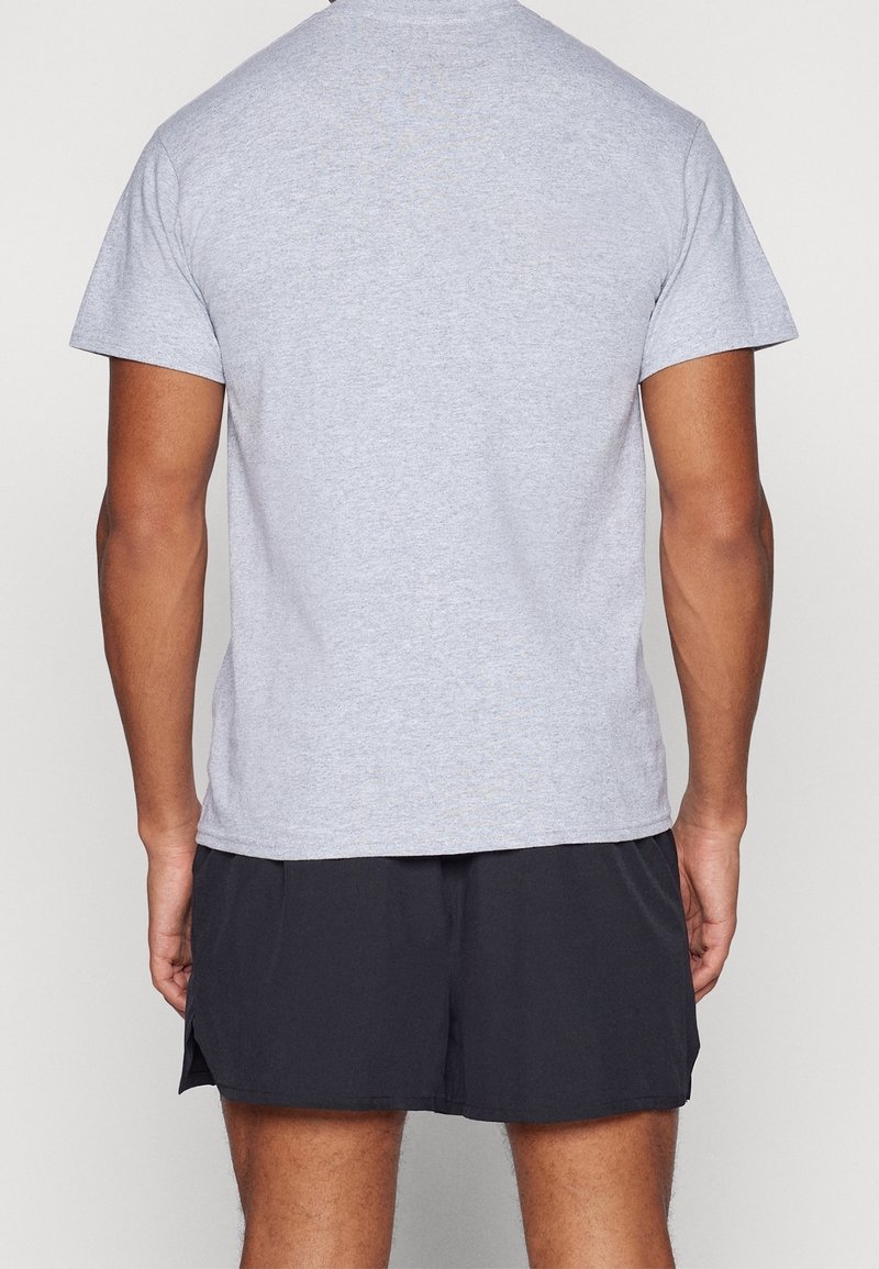Hombre vistiendo una camiseta de manga corta gris claro y pantalones cortos deportivos negros, visto desde atrás contra un fondo sencillo.