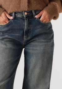 Hoge taille donkere denim jeans met een licht vervaagde afwerking, met voorzakken en knopen, gecombineerd met een gestructureerde bruine trui.